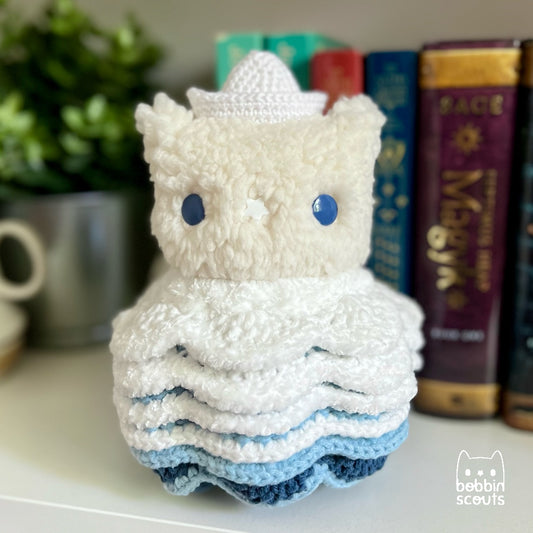 cream plushie with a crocheted shell made to look like ocean waves and a tiny sailboat on its head